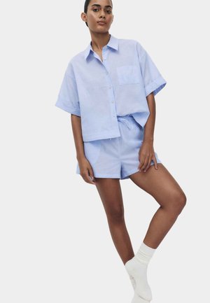 Joven mujer con camisa de manga corta azul claro abotonada y pantalones cortos a juego, posando con una pierna ligeramente levantada, con calcetas blancas.
