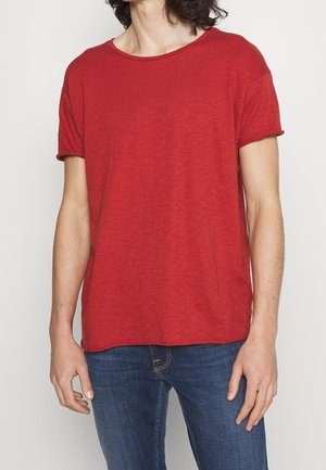 Osoba ubrana w zwykły czerwony t-shirt z krótkim rękawem i niebieskie dżinsy, stojąca na jasnym tle z luźno opuszczonymi rękami wzdłuż ciała.