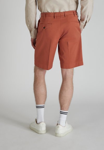 Shorts color ruggine in cotone, con una parte davanti liscia, tasche laterali e posteriori, e una leggera lucentezza nella trama del tessuto. Indossati con scarpe da ginnastica bianche.