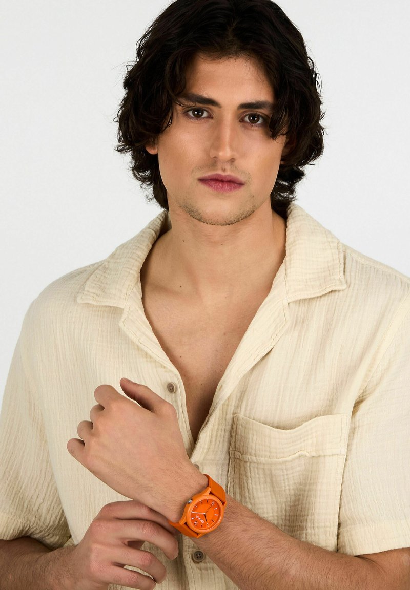 Orologio in silicone arancione con quadrante circolare e indici delle ore in grassetto, indossato da una persona con una camicia beige texturizzata.