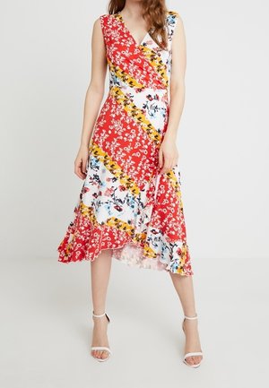 Vrouw met een mouwloze, V-hals, kuitlange jurk met rode, gele en witte bloemenpatronen en witte sandalen met hoge hakken.