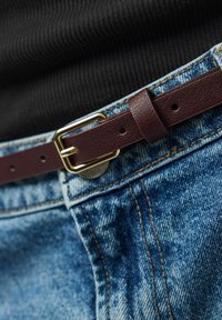 Gros plan d'une ceinture en cuir marron passée dans la boucle d'un jean en denim bleu porté avec un haut noir côtelé.