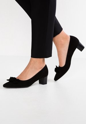 Femme portant des chaussures noires en daim à talons blocs bas avec un détail nœud, associées à un pantalon noir sur fond blanc.