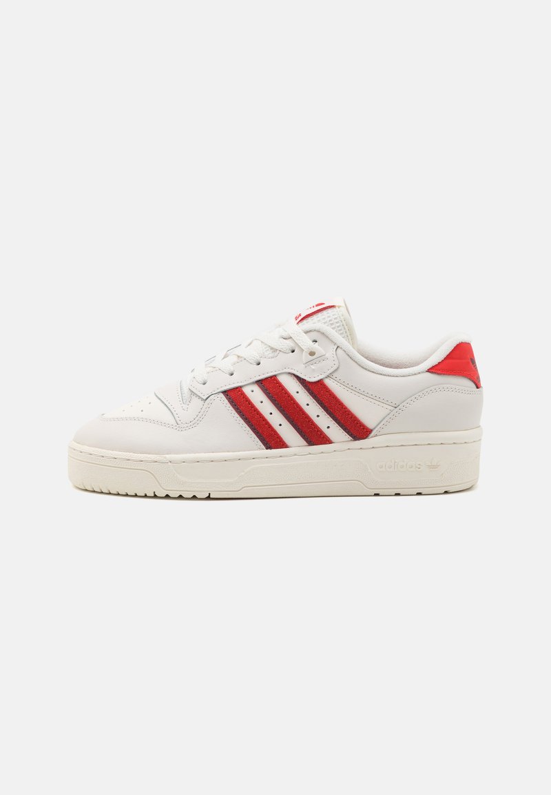 Biele kožené tenisky s červenými semišovými pruhmi a červenými akcentmi. Zaoblená špička, šnurovacie uzavretie a textúrovaná gumová podrážka. Logo Adidas na päte.