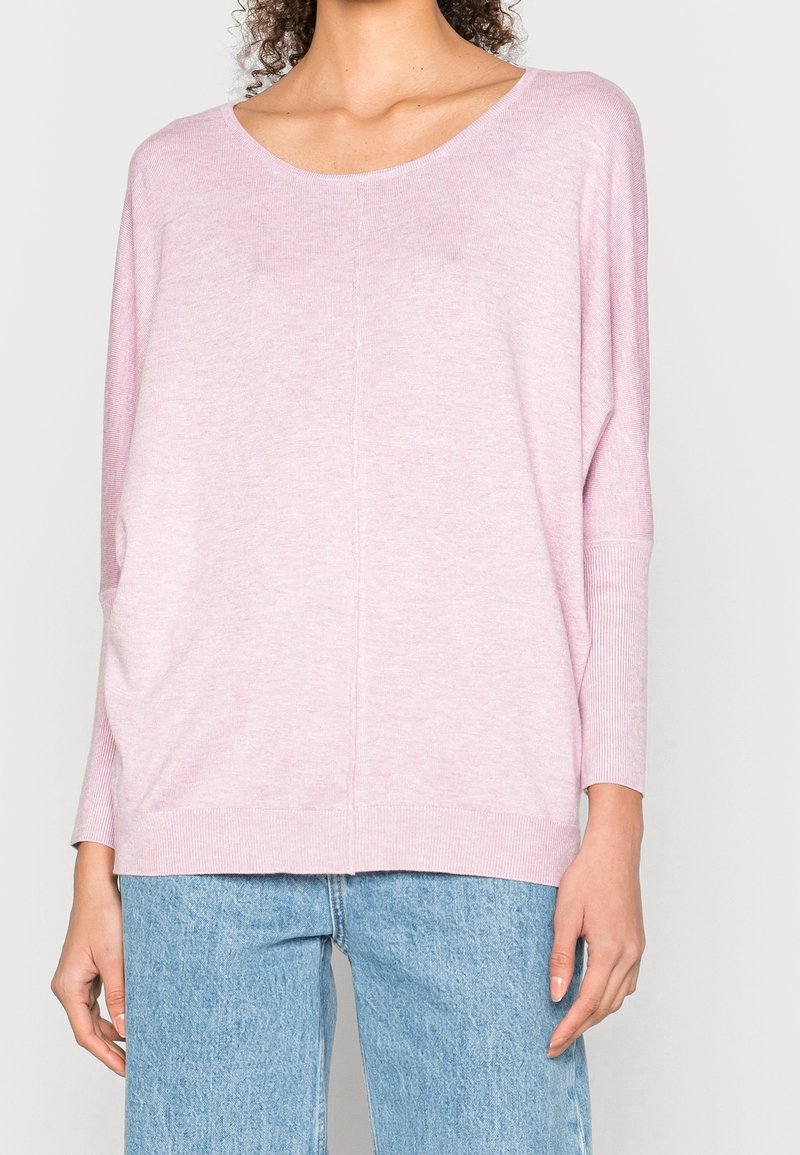 Femme portant un pull ample à manches longues rose clair avec un large col et un jean en denim bleu clair, cadré au-dessus du cou.