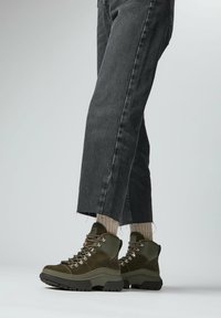 Olijfgroene wandelboots met een bovenwerk van suede en leer, vetersluiting, robuute zool en textuurelementen, gecombineerd met grijze denim.