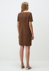 Bruine suède jurk met korte mouwen, rechte snit en een achternaad. Model draagt neutraalgekleurde sleehakken. Minimalistisch ontwerp.