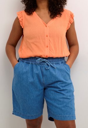 Frau trägt eine orange ärmellose Bluse mit Rüschenärmeln und blaue Jeans-Shorts mit Kordelzug, die Hände in den Taschen.