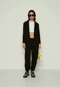 Γυναίκα με μαύρο φαρδύ σακάκι και cargo παντελόνι, λευκή cropped μπλούζα, μαύρα γυαλιά ηλίου, μαύρα και άσπρα sneakers, κρατώντας μικρή κίτρινη τσάντα.