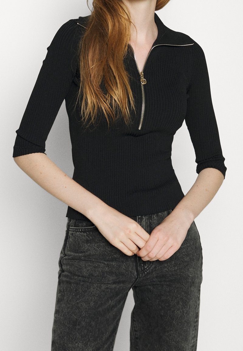 Zwart geribbeld shirt met een halve ritssluiting bij de kraag, driekwart mouwen en een aansluitend model. Model draagt het met vervaagde zwarte jeans.