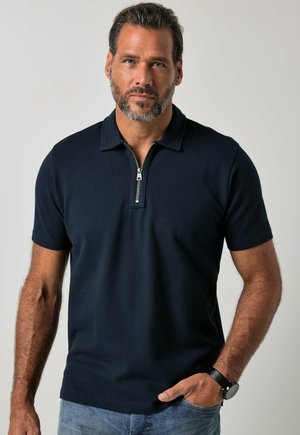 Uomo con capelli corti e scuri e barba, che indossa una polo blu navy scuro con zip al collo e jeans azzurri chiari su sfondo grigio.