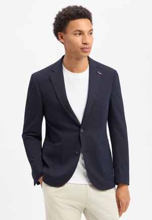 Junger Mann trägt ein marineblaues Sakko über einem weißen T-Shirt und beige Hose, steht mit den Händen in den Taschen und schaut leicht nach rechts.