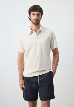 Hombre con cabello rizado oscuro, vestido con una camisa polo de manga corta color crema y pantalones cortos negros con cordón, de pie frente a un fondo liso.