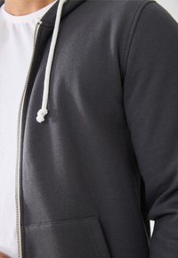 Close-up of a person wearing a dark gray zip-up hoodie with white drawstrings over a white shirt.