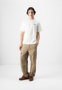 White cotton t-shirt with blue "Timberland" logo, paired with beige cargo pants featuring large pockets and a relaxed fit. Brown shoes complete the look.