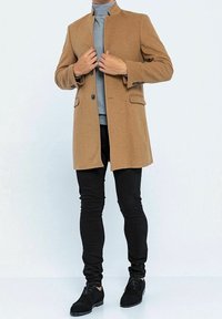 Cappotto in misto lana marrone con colletto alto, chiusura a un bottone e due tasche frontali, abbinato a un dolcevita grigio chiaro e pantaloni neri.