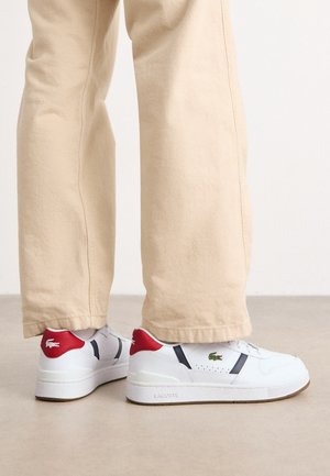 Zapatillas blancas con acentos en rojo y azul marino, que presentan un diseño perforado y un logo verde. Suela de goma texturizada y líneas limpias.