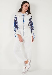 Blusa bianca con ricami floreali blu e frange, abbinata a pantaloni bianchi a gamba larga e scarpe da ginnastica bianche.