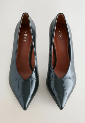 Un par de zapatos planos de cuero negros con punta afilada y un patrón bordado, con plantillas marrones de la marca "NEXT", mostrados desde arriba sobre un fondo claro.