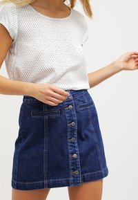 Bílý top s krátkým rukávem a černým tečkovaným vzorem, v kombinaci s modrou džínovou sukní s předními knoflíky a kapsami, strukturovaná látka.