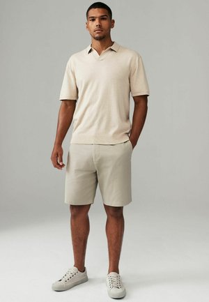 Camisa polo beige de manga corta con un diseño de cuello en V, combinada con pantalones cortos de caqui claro y zapatillas gris claro. Textura de tela suave.