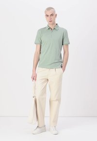 Svetlo zelena polo majica z zadrgo na ovratniku, v kombinaciji s kremnimi hlačami in belimi supergami. Ima enostaven dizajn in mehak material.