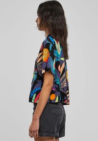 Camicetta nera a maniche corte, morbida, con motivi floreali colorati. Indossata con pantaloni corti grigi casual. Materiale in cotone, design rilassato.