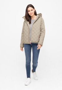 Gequiltde olijfgroene jas met een capuchon, ritssluiting en roze accent. Draagt over een gestreept topje en gecombineerd met blauwe spijkerbroeken en witte sneakers.