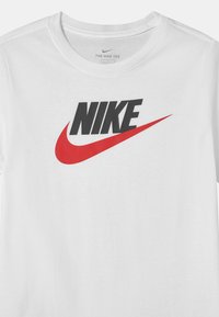 Camiseta de algodón blanca con cuello redondo, que presenta un logotipo negro de "NIKE" y un gráfico de swoosh rojo impresos en la parte delantera.