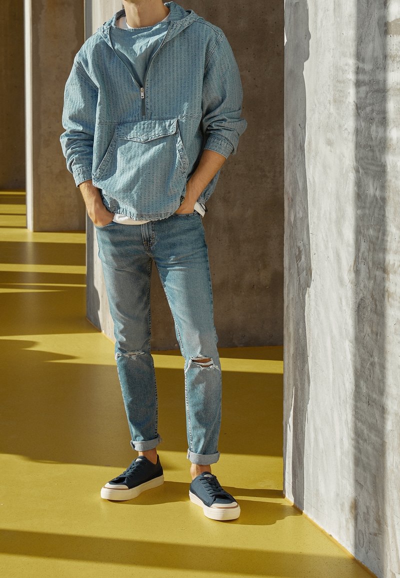 Chaqueta azul claro con capucha y cremallera, con patrón texturizado, bolsillo delantero, combinada con vaqueros azules desgastados y zapatillas oscuras sobre un suelo amarillo.