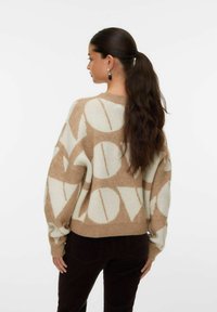 Pull en crème et beige à motifs avec une coupe surdimensionnée, présentant des formes abstraites, un col rond et un ourlet côtelé. Porté avec un pantalon marron foncé.