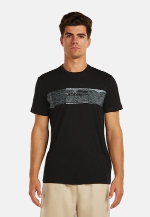 Mężczyzna ubrany w czarną koszulkę z krótkim rękawem z poziomym abstrakcyjnym szarym wzorem na klatce piersiowej oraz beżowe spodnie, stojący na jednolitym tle.