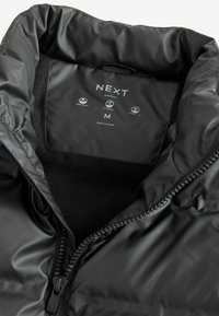 Chaqueta acolchada negra con una textura exterior suave, forro de felpa, cierre de cremallera y etiqueta de cuidado que muestra características impermeables y tamaño M.
