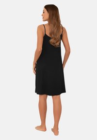 Kobieta z długimi falującymi włosami, ubrana w bezrękawową czarną sukienkę do kolan, stoi boso tyłem na jednolitym jasnym tle.