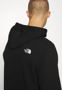 Black hoodie with a kangaroo pocket, features a white logo on the left shoulder. Material appears soft and textured, with a double-layered hood.
