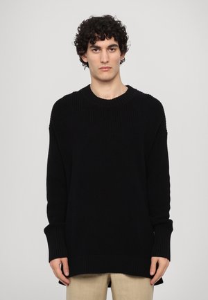 Young man with curly hair wearing an oversized black knit sweater and beige pants, standing against a plain light background.