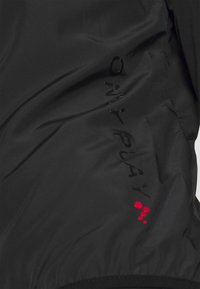 Zwarte stoffen jas met een soepele textuur. Heeft een glanzend zwart "ONLY PLAY" logo en rode stipjes als accent. Lichtgewicht ontwerp.