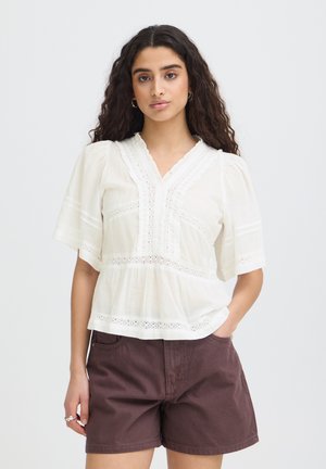 Kvinde med langt krøllet hår iført en hvid bluse med blonder og brune shorts, står med den ene hånd i lommen.