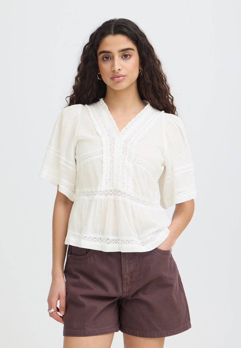 Femme aux longs cheveux bouclés portant un chemisier blanc bordé de dentelle et un short marron, debout avec une main dans la poche.