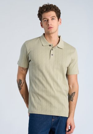 Junger Mann mit lockigem Haar, der ein beiges kurzärmeliges Poloshirt und blaue Jeans trägt, zeigt Tätowierungen eines Adlers und eines Schmetterlings auf den Unterarmen.