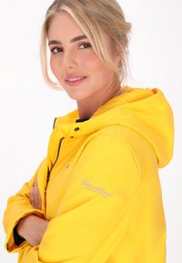 Gul vattentät jacka med huva, med dragkedja fram och logotyp på ärmen. Slät textur, något figursydd design.