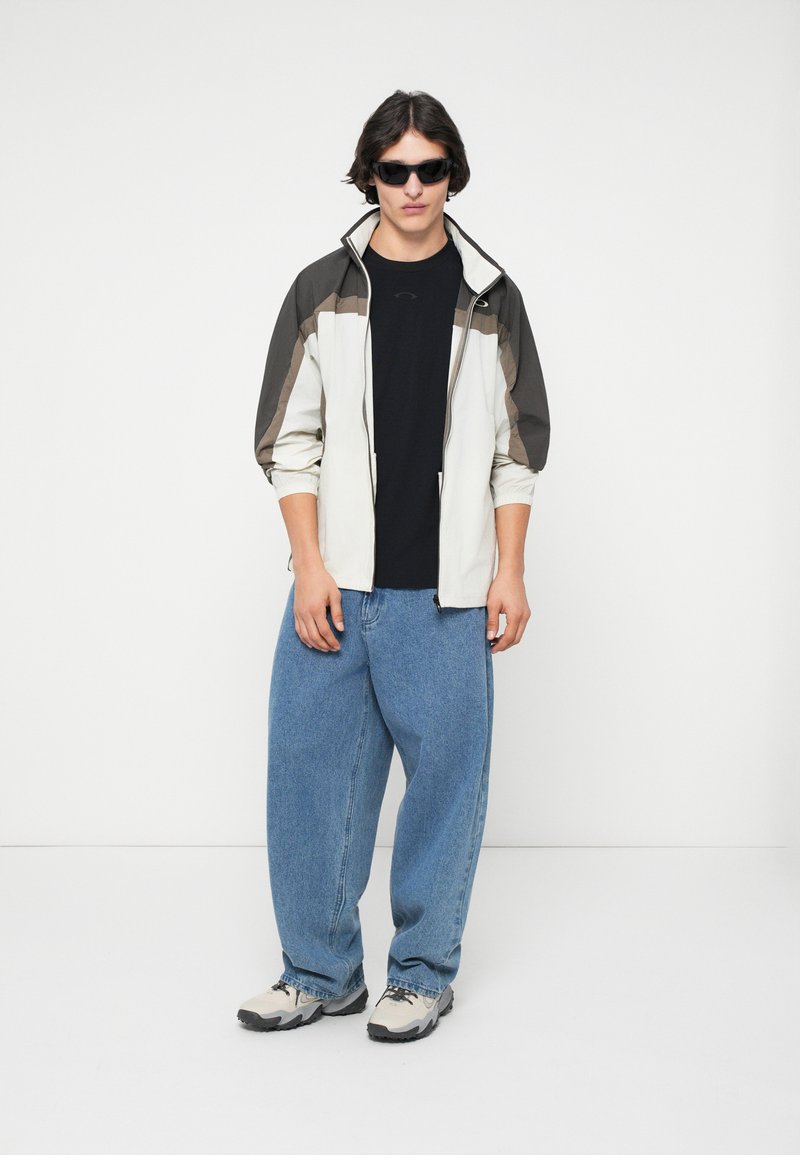 Veste zippée légère crème avec des accents gris foncé, assortie à un jean bleu ample et des chaussures de sport grises. T-shirt noir en dessous.