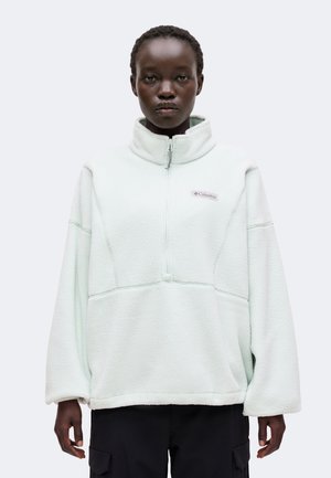 Personne portant un pull polaire blanc Columbia avec une fermeture éclair sur la moitié et un pantalon noir, debout contre un fond clair uni.