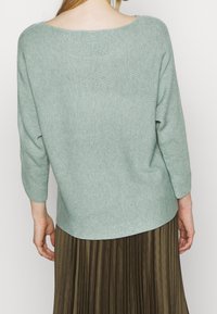 Lichtgroene, geribbelde trui met brede ronde hals en driekwart mouwen, gecombineerd met een geplooide bruine rok. Textuur van de stof is zichtbaar.