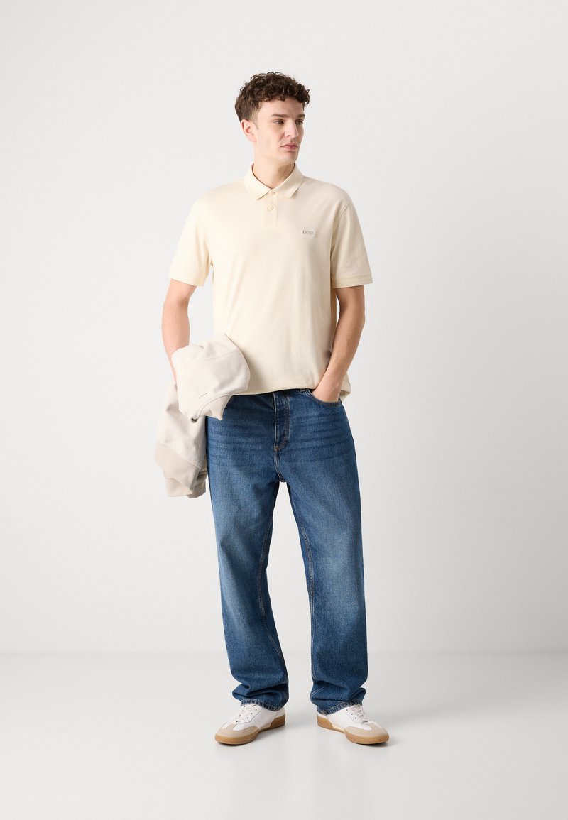 Polo shirt beige clair, jeans bleus décontractés et baskets blanches avec des accents beiges. Sweatshirt gris supplémentaire tenu à la main gauche. Design minimaliste.