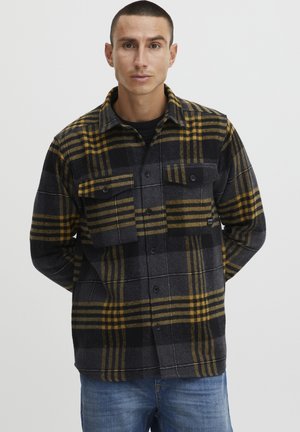 Camisa de franela a cuadros en negro y amarillo con dos bolsillos frontales, cierre de botones y cuello, con una apariencia texturizada similar a la lana.