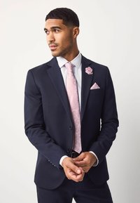 Sötétkék öltöny texturált felülettel, világos rózsaszín nyakkendővel és zsebkendővel, virágmintával. Rózsaszín kitűző a hajtókán.