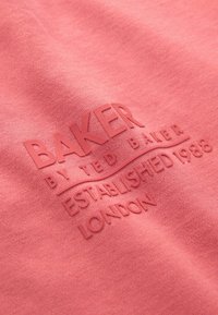 Roze stof met reliëflogo "BAKER by TED BAKER" en "OPGERICHT IN 1988 LONDEN" voor een textuur met verhoogd detail.