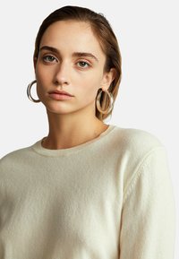 Suéter crema hecho de una tela suave, con un escote redondo y ribetes acanalados, combinado con grandes pendientes de aro plateados.