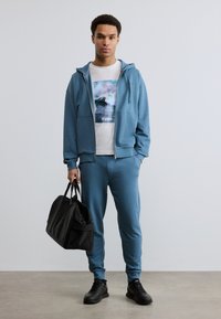 Νέος άνδρας με μπλε φόρμα και λευκό T-shirt με σχέδιο, κρατώντας μια μαύρη τσάντα duffel, στέκεται μπροστά σε απλό φωτεινό τοίχο.